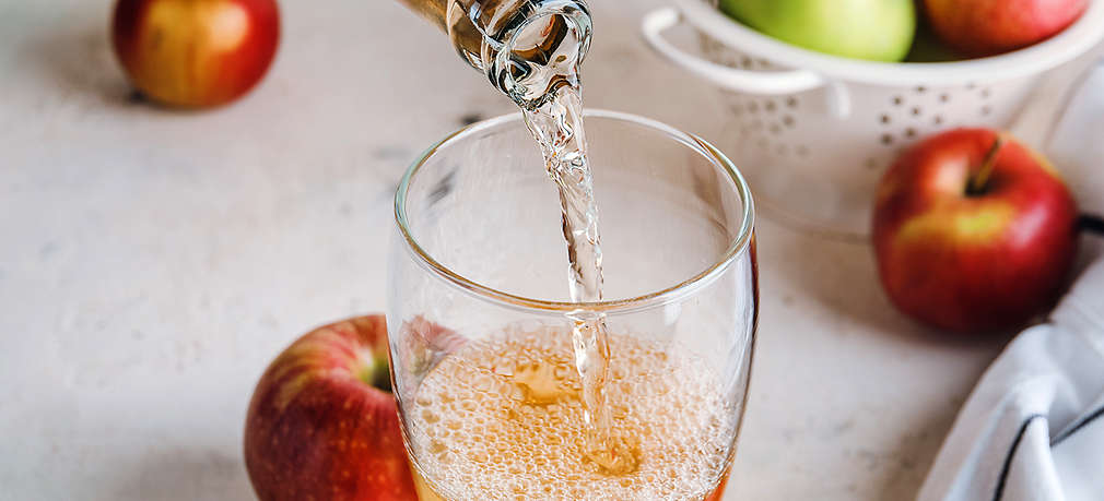 Cidre wird in ein Glas geleert, daneben liegen Äpfel auf dem Tisch sowie in einem weißen Sieb