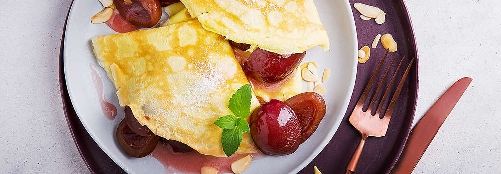 Zwei zusammengefaltete, geviertelte, dünne Pfannkuchen auf einem Teller mit Soße, eingekochten Pflaumen, Zitronenmelisse und Mandelblättchen serviert