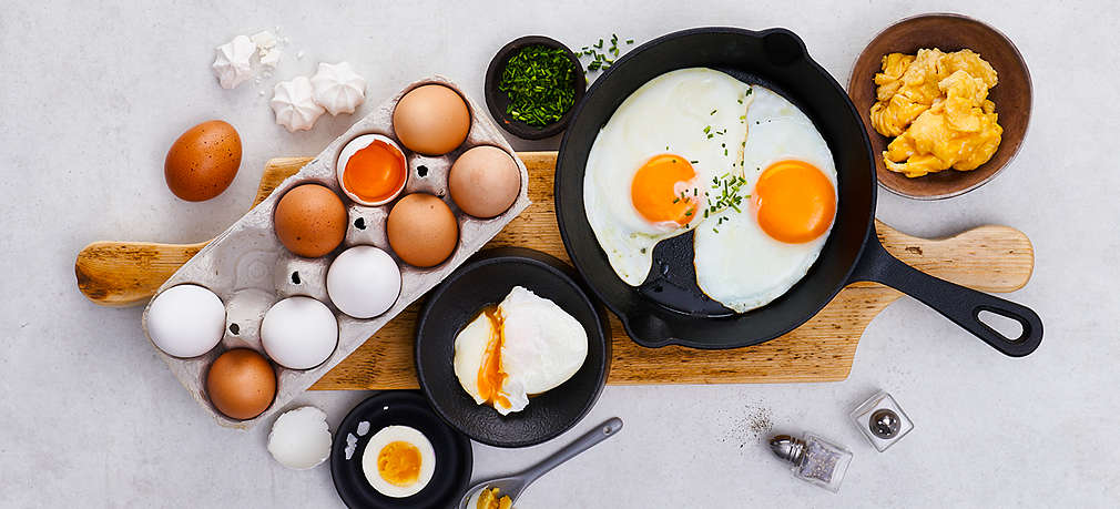 Rohe Eier in Verpackung, daneben Spiegelei in Bratpfanne, pochierte Eier in Teller, Omelett in kleiner Schale sowie geschnittener Lauch und Salz- und Pfefferstreuer. 
