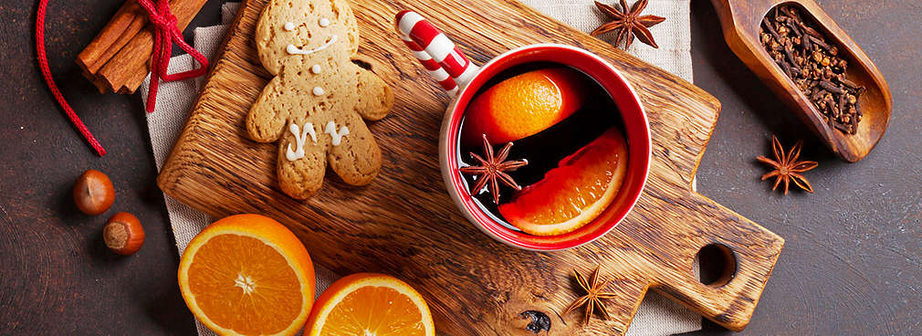 Eine Tasse Glühwein angerichtet auf einem Holzbrett neben einem Lebkuchenmännchen, frischen Orangen und unterschiedlichen weihnachtlichen Gewürzen