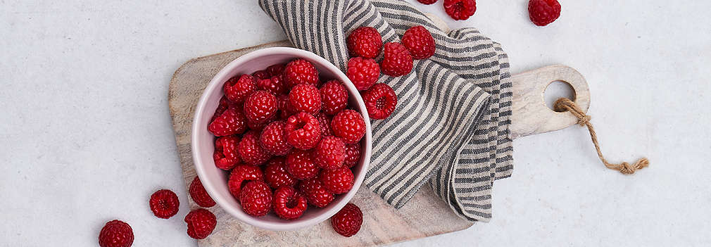 Eine Schale mit Himbeeren steht auf einem Schneidebrett auf einem Tisch. Daneben liegt ein Küchentuch. 