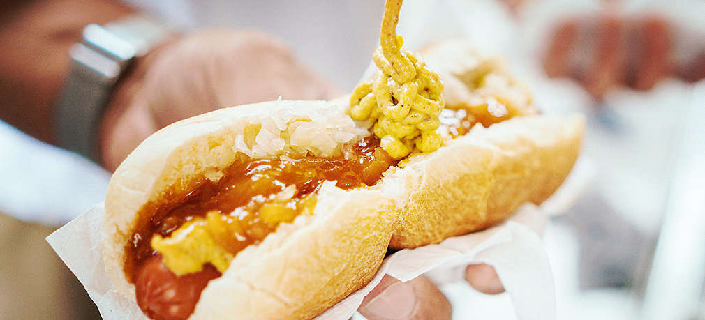 Eine Hand hält einen frischen Hot Dog mit Würstchen und Zwiebelsauce, während die andere Hand den Hot Dog mit Senf aus der Tube toppt
