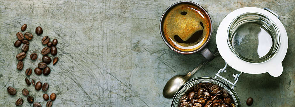 Mehrere Kaffebohnen liegen auf dem Tisch. Ihnen gegenüber steht eine Tasse Kaffee und ein geöffnetes Glas mit Kaffeebohnen.