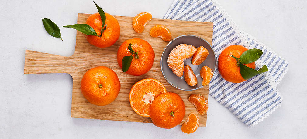 Mehrere Mandarinen liegen auf einem Holz-Schneidebrett auf einem Tisch. Eine davon ist halbiert und eine andere geschält.