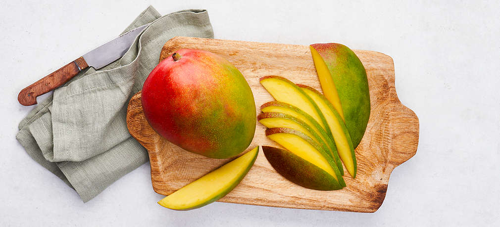 Eine ganze Mango-Frucht liegt auf einem Holzschneidebrett. Links daneben liegt ein Küchenmesser auf einem Küchentuch und rechts auf dem Schneidebrett liegt eine Hälfte einer Mangofrucht in Stücke geschnitten.