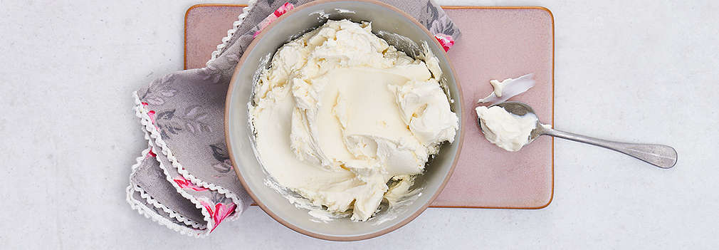 Eine mittelgroße Schale mit Mascarpone gefüllt steht auf einem Tisch. Daneben liegt ein Esslöffel.