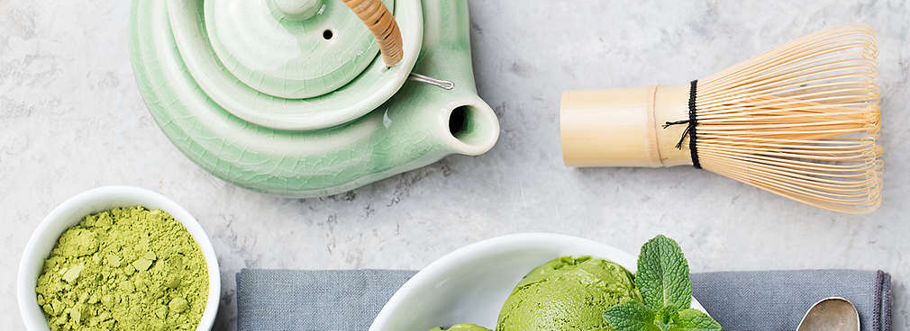 Eine kleine Kanne Matcha-Tee steht auf einem Tisch. Daneben steht eine kleine Schale mit Matcha-Tee Pulver, eine Schale mit Matcha-Eis und einem Matcha-Besen.