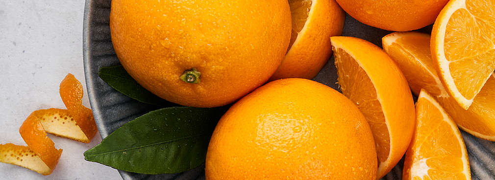 Zwei ganze Orangen, Orangenschnitze sowie Orangenzeste liegen auf einem Teller