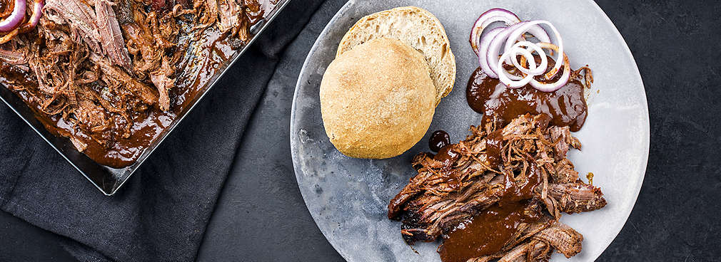 Pulled Pork angerichtet auf einem Teller neben einem Burger-Brötchen und roten Zwiebeln