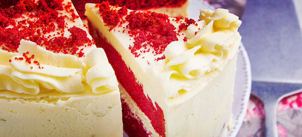 Abbildung eines Red-Velvet-Kuchens im Closeup auf einem Teller, von dem ein großes Stück ausgeschnitten, das leicht herausgezogen ist, sodass man die inneren zwei roten Schichten und die weiße Schicht in der Mitte sehen kann. Rechts neben dem Kuchenteller liegt eine gemusterte Serviette und darauf ein Tortenheber