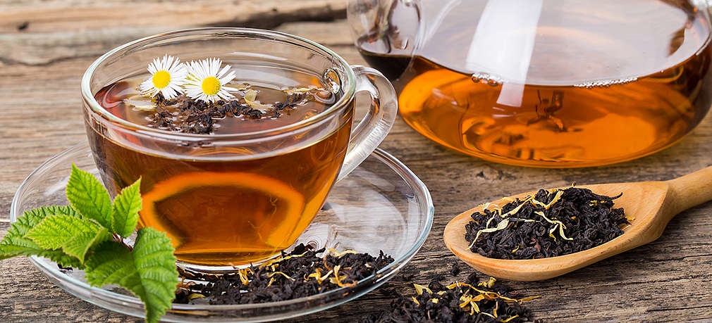 Schwarzer Tee in einer Tasse und einer Teekanne auf Holzuntergrund, rechts daneben liegt ein Löffel mit losem schwarzen Tee 