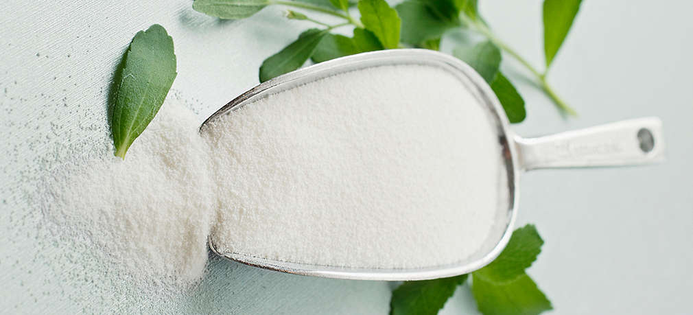 Stevia in Pulverform in Dosierschaufel und daneben frische Steviablätter