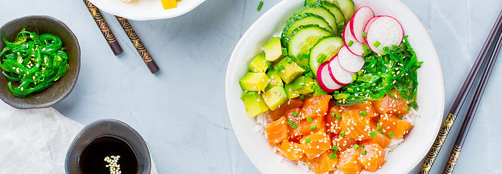 Zu sehen ist eine Sushi Bowl mit Reis, rohen Lachswürfeln, Avocadowürfeln, Gurkenscheiben, Radieschenscheiben und Seetang. Eine weitere Schale ist links im Bild, darin sind Reis, Tofuwürfel, Mangowürfel, rote Zwiebelscheiben, Avocadowürfel und Radieschenscheiben. Außerdem im Bild: zwei Paar Stäbchen und drei Schälchen mit Schnittlauch, Seetang und Sojasoße