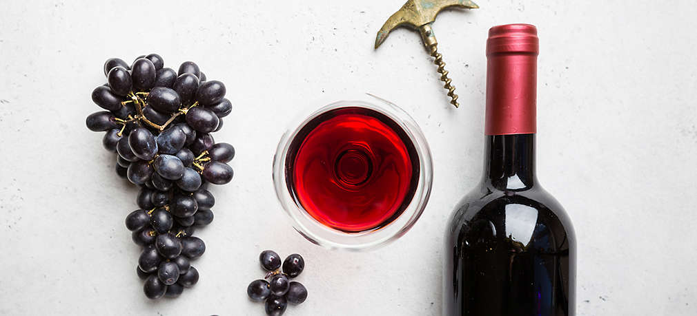 Eine Rispe roter Weintrauben, daneben ein Glas Rotwein sowie eine Rotweinflasche und ein Flaschenöffner von oben fotografiert auf hellgrauem Grund