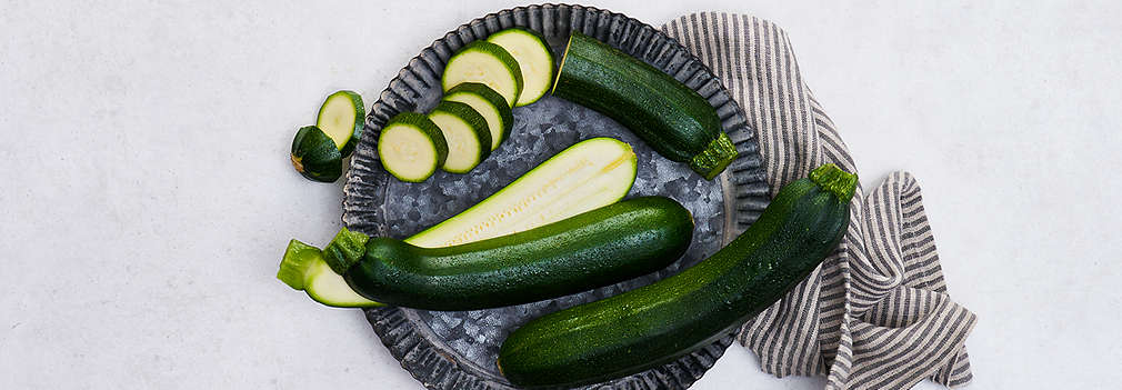 Zwei grüne angeschnittene und eine ganze Zucchini liegen auf einem Teller. 