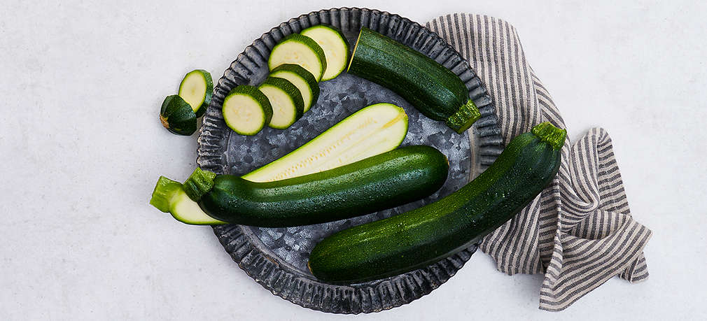 Zwei grüne angeschnittene und eine ganze Zucchini liegen auf einem Teller. 