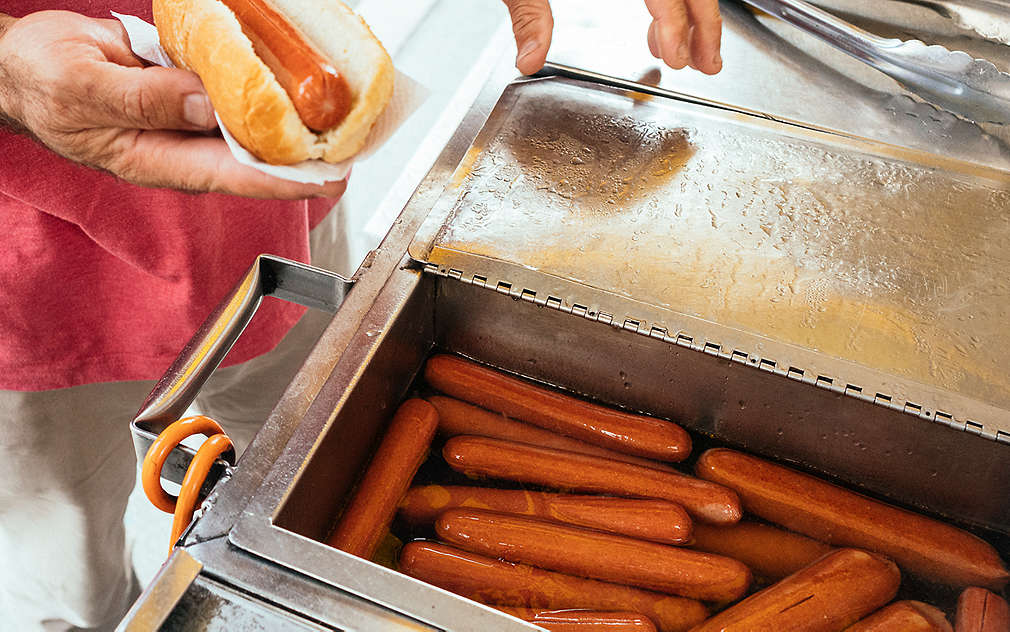 Na stoisku z hot dogami stoją metalowe kotły z gotowanymi parówkami pływającymi w wodzie.