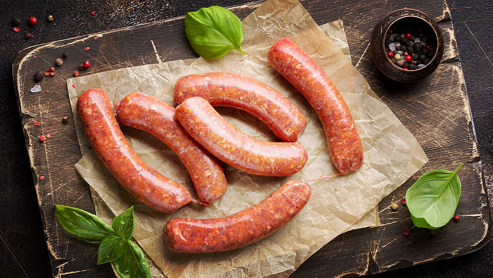 Показани са наденички Merguez, гарнирани с босилек и цветни чушки върху дървена дъска.