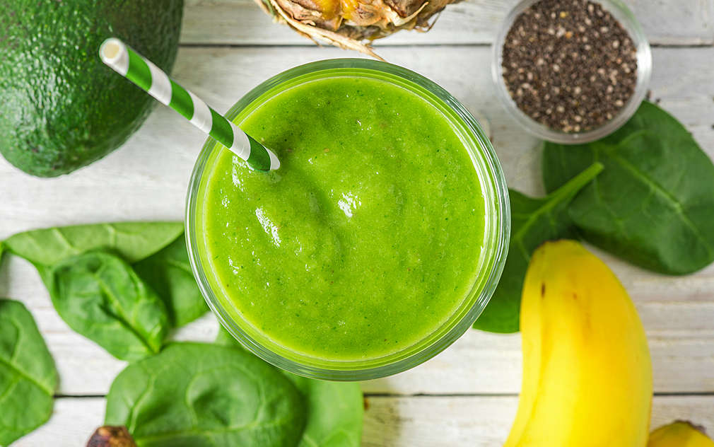 Iz ptičje perspektive vidi se zeleni smoothie u čaši na drvenoj površini. Uokolo se nalaze avokado, listovi špinata, banane, zdjelica s chia sjemenkama i ananas.