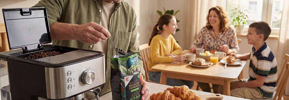 Ein Mann füllt Espressobohnen aus einer K-Gold Packung in eine Kaffeemaschine, während im Hintergrund eine Frau und zwei Kinder gemeinsam an einem gedeckten Frühstückstisch sitzen., Ein Mann f�llt Espressobohnen aus einer K-Gold Packung in eine Kaffeemaschine, w�hrend im Hintergrund eine Frau und zwei Kinder gemeinsam an einem gedeckten Fr�hst�ckstisch sitzen.