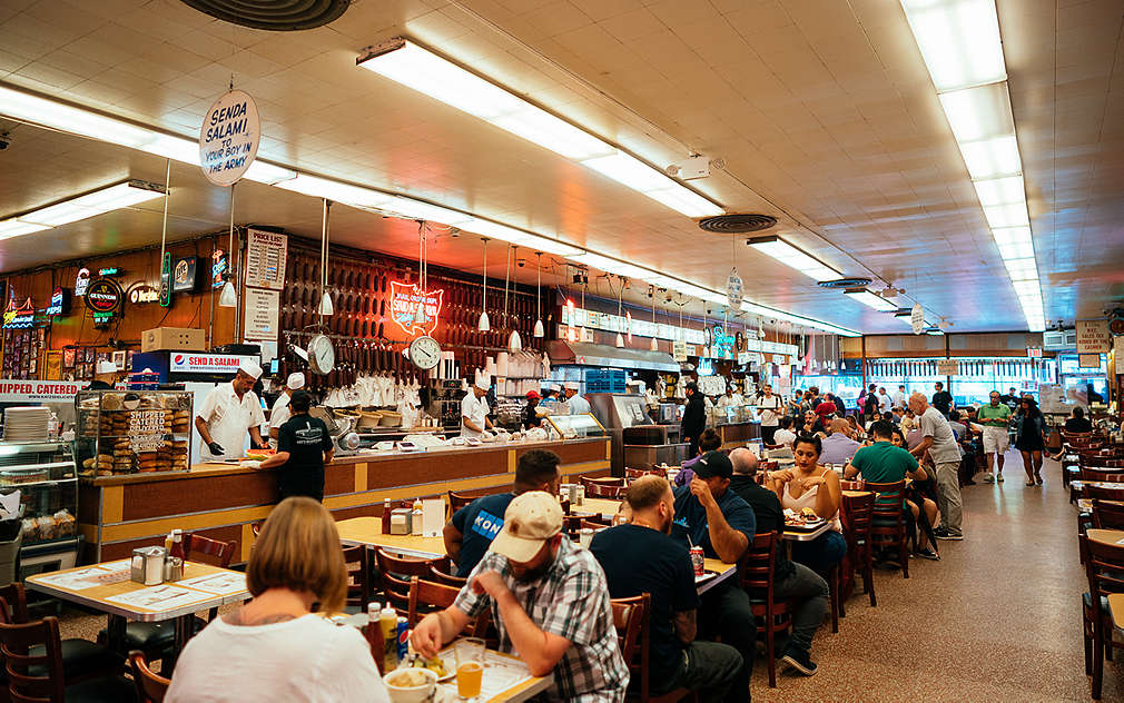 Zdjęcie wnętrza Katz's Deli w Nowym Jorku z barem i kilkoma rzędami stołów z krzesłami w stylu retro, przy stołach widać jedzących ludzi.