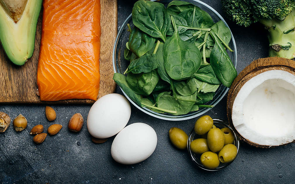Na tamnoj radnoj površini nalaze se različite svježe namirnice kao što su losos, sir, jaja, orašasti plodovi, kokos, masline, avokado i zeleno povrće.