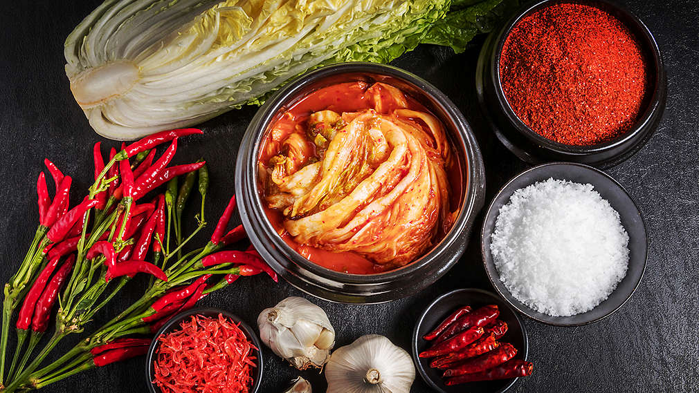 O suprafață de lucru închisă la culoare, fotografiată de sus, pe care se pot vedea varză chinezească, usturoi și boluri mici cu pudră de chili, semințe de chili și sare de mare. În mijloc se poate vedea un bol mare plin cu kimchi.