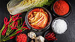 Seshora vyfocená tmavá pracovní plocha, na které leží čínské zelí, malé svazečky chilli, česnek a malé mističky s mletým chilli, chilli papričkami a mořskou solí. Uprostřed je větší mistička plná kimchi.