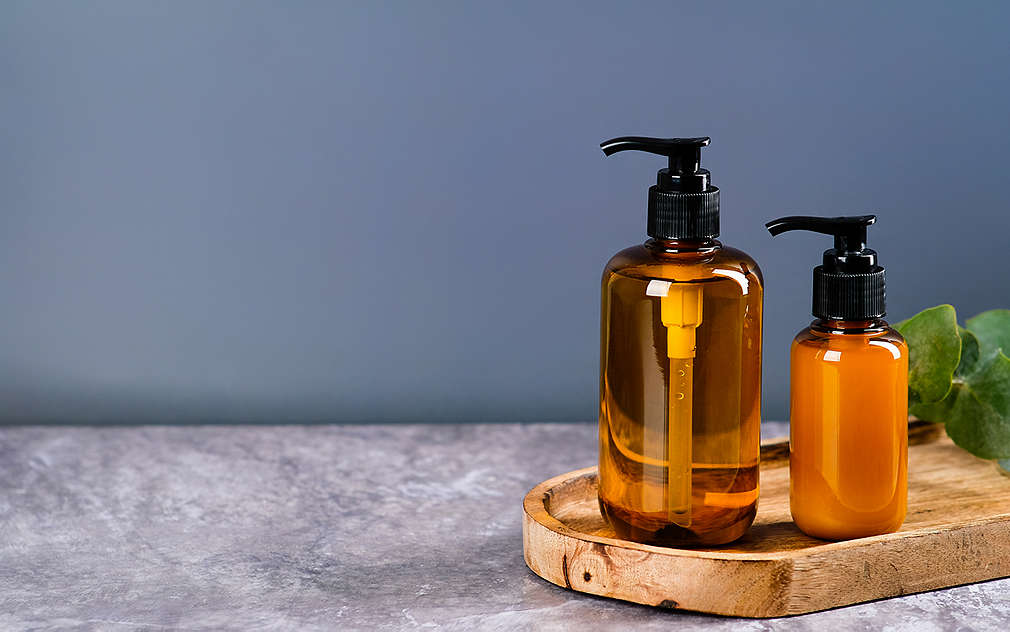  Вижда се овална дървена дъска на сив мраморен фон, върху която са поставени два кафяви дозатора за сапун, пълни с прозрачен и млечен сапун, сиво-син фон.