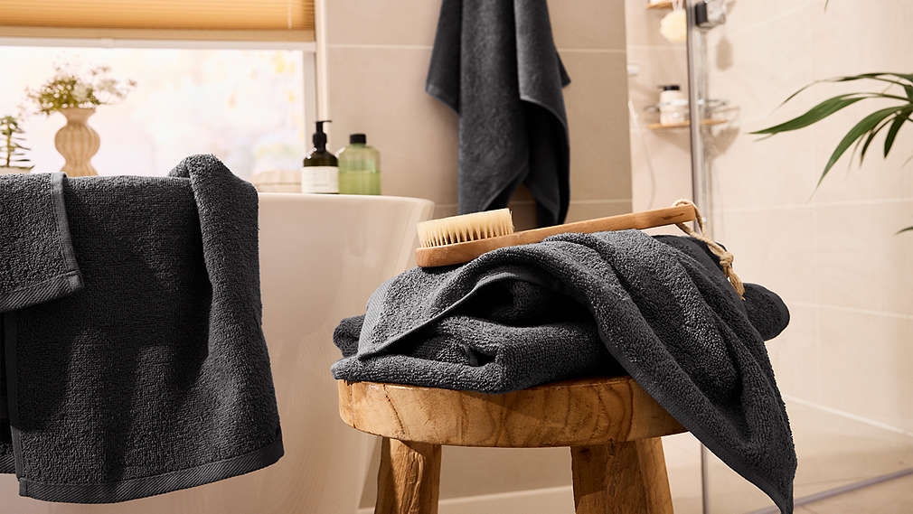 Dunkelgraue, saugfähige Handtücher von LIVARNO liegen dekorativ auf einem Holzhocker und einer Badewanne bereit, um für wohnliche Gemütlichkeit im Bad zu sorgen., Dunkelgraue, saugf�hige Handt�cher von LIVARNO liegen dekorativ auf einem Holzhocker und einer Badewanne bereit, um f�r wohnliche Gem�tlichkeit im Bad zu sorgen.