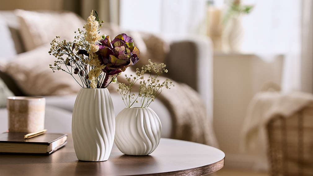 Zwei stilvoll geformte weiße Vasen von LIVARNO mit Trockenblumen stehen als dekorative Accessoires auf einem Couchtisch in einem hellen Wohnzimmer., Zwei stilvoll geformte wei�e Vasen von LIVARNO mit Trockenblumen stehen als dekorative Accessoires auf einem Couchtisch in einem hellen Wohnzimmer.