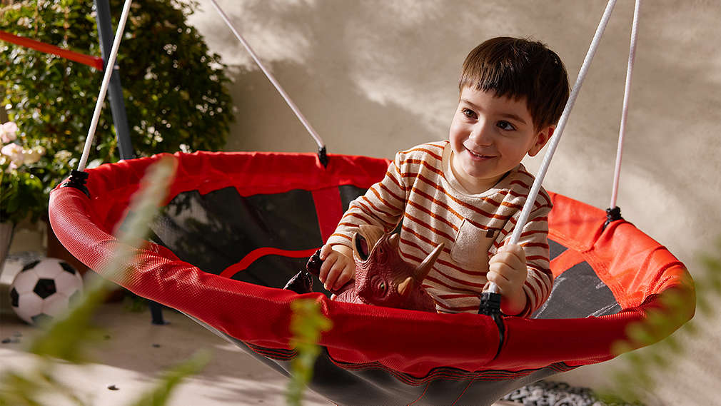 Ein lächelndes Kind in gestreifter Lupilu Kinderkleidung sitzt mit einem Spielzeug-Dinosaurier auf einer rot-schwarzen Schaukel im Freien., Ein l�chelndes Kind in gestreifter Lupilu Kinderkleidung sitzt mit einem Spielzeug-Dinosaurier auf einer rot-schwarzen Schaukel im Freien.