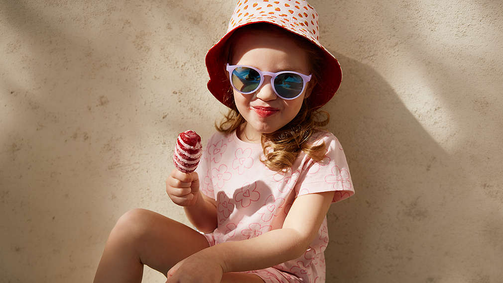 Ein Kind mit Sonnenhut und Sonnenbrille sitzt entspannt vor einer hellen Wand und hält ein rot-weiß gestreiftes Wassereis in der Hand., Ein Kind mit Sonnenhut und Sonnenbrille sitzt entspannt vor einer hellen Wand und h�lt ein rot-wei� gestreiftes Wassereis in der Hand.