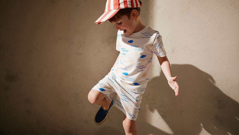 Ein kleiner Junge trägt ein sommerliches Set mit Fisch-Motiven und dazu passende, dunkelblaue Lupilu Kinderschuhe., Ein kleiner Junge tr�gt ein sommerliches Set mit Fisch-Motiven und dazu passende, dunkelblaue Lupilu Kinderschuhe.