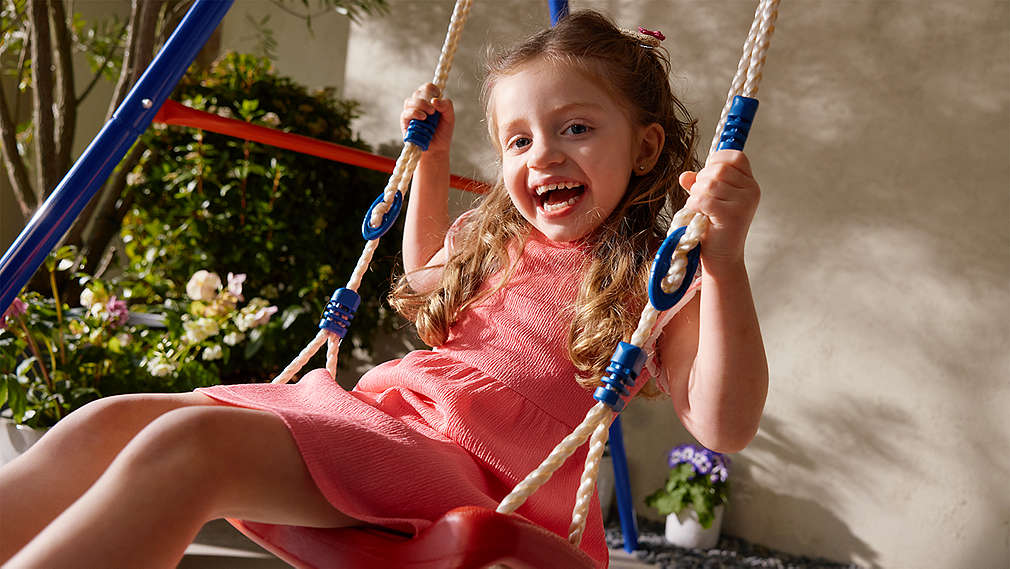 Ein lachendes Kind in einem hochwertigen korallenfarbenen Kleid hält sich sicher an den Seilen einer Schaukel fest., Ein lachendes Kind in einem hochwertigen korallenfarbenen Kleid h�lt sich sicher an den Seilen einer Schaukel fest.