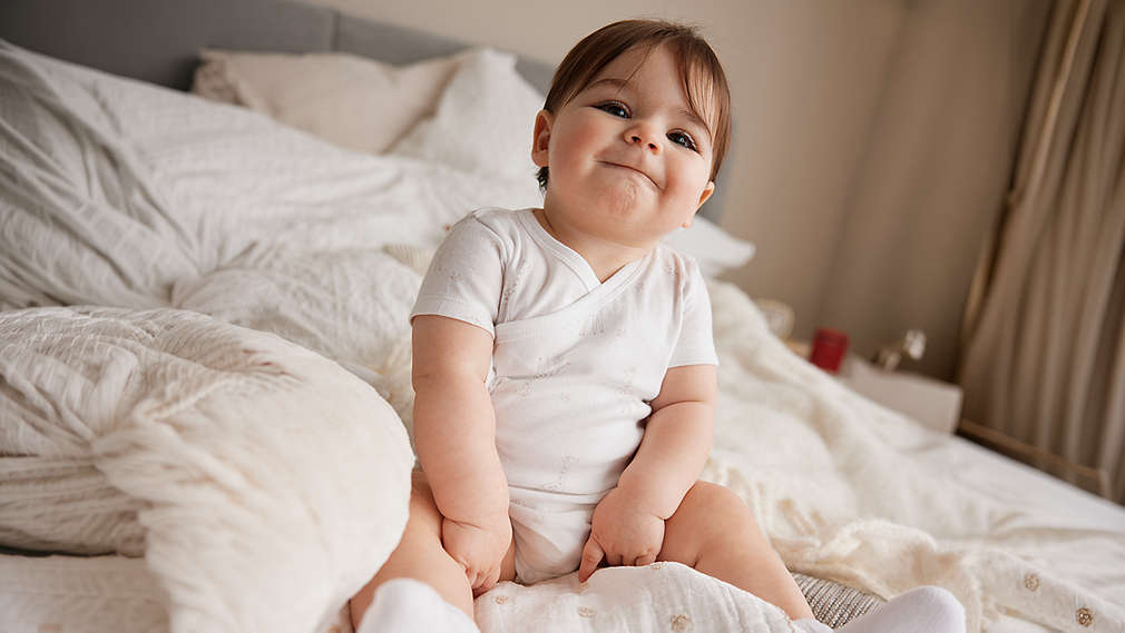 Ein zufrieden schauendes Baby sitzt in einem weißen Wickelbody auf einer gemütlichen, hellen Bettlandschaft., Ein zufrieden schauendes Baby sitzt in einem wei�en Wickelbody auf einer gem�tlichen, hellen Bettlandschaft.