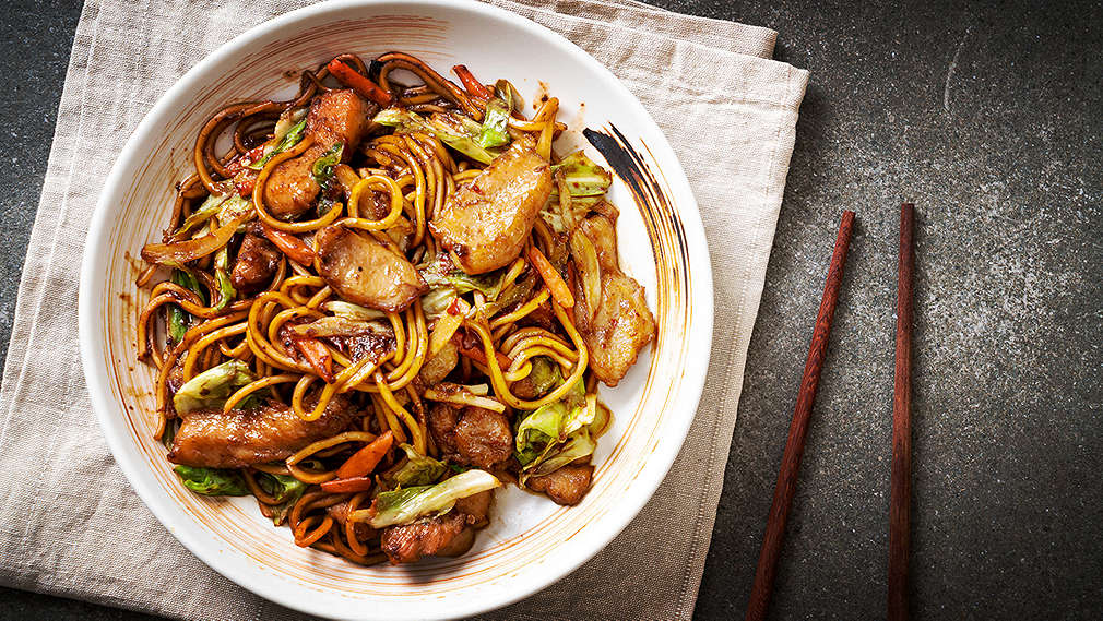A white plate on a napkin with grey background. On the plate, you can see an asian dish with meat and noodles.