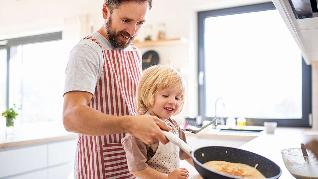 Na obrázku je muž, ktorý drží v ruke panvicu. Stojí v kuchyni s dieťaťom, obaja majú oblečené zástery a pripravujú palacinky.