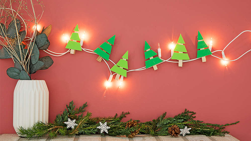 Na ružovej stene visí svietiaca LED svetelná reťaz. Je dozdobená vlastnoručne vyrobenými vianočnými stromčekmi. Vedľa nej stojí biela váza so stonkami.