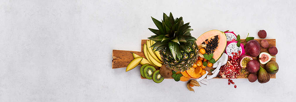 Доска с богатым выбором экзотических фруктов, например ананас, папайя и питахайя на светло-сером фоне.