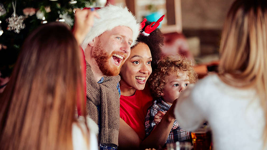 Skupina ľudí, v centre pozornosti mladý muž, mladá žena a malé dieťa. Muž má na hlave mikulášsku čiapku, žena tiež vianočnú čelenku a v pozadí je vidieť ozdobený vianočný stromček.