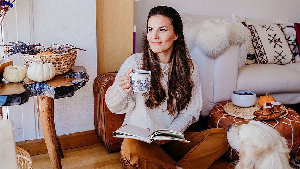 Na obrázku je mladá žena v džínsoch a vo svetlom pletenom svetri, sedí v tureckom sede na pohodlnej pohovke v obývačke. Číta knihu a v jednej ruke drží šálku čaju.