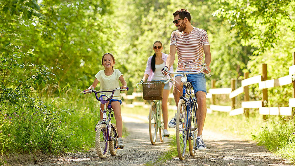 Na obrázku je žena, muž a dieťa, ktorí idú pri slnečnom počasí na bicykloch po lesnej ceste.