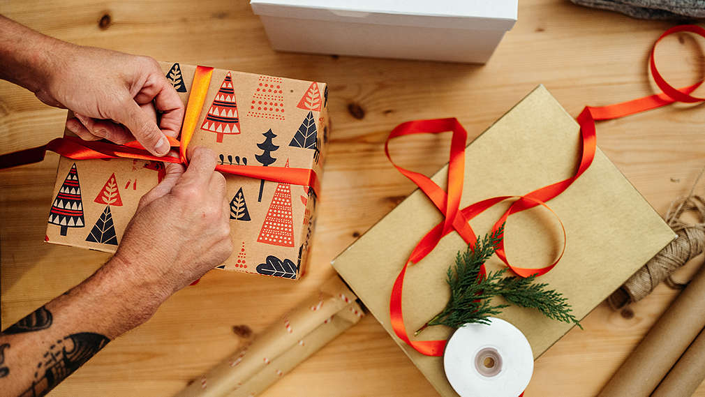 Mužské ruky balia vianočné darčeky na stole s vianočným papierom, stuhami a vetvičkou.
