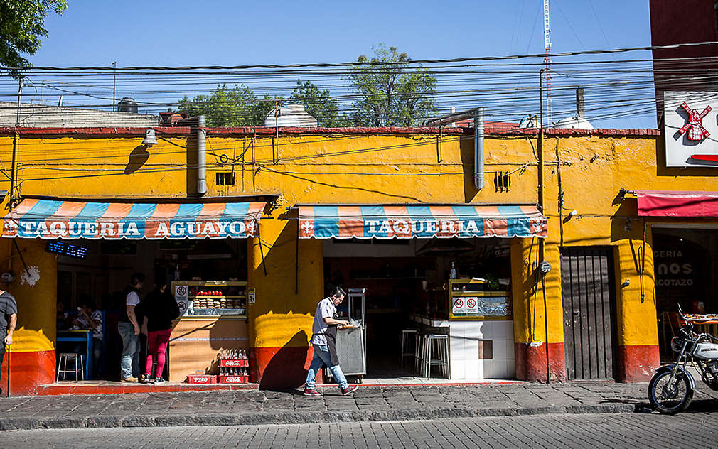 Na obrázku je mexická taquería se žlutou fasádou a modro-oranžově pruhovanou markýzou.
