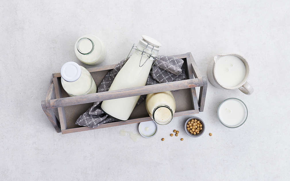 Različiti mliječni proizvodi iz ptičje perspektive u čašama i bocama na svijetloj površini, djelomično i u drvenoj kutiji s ručkom u kojoj se nalazi i siva kuhinjska krpa.
