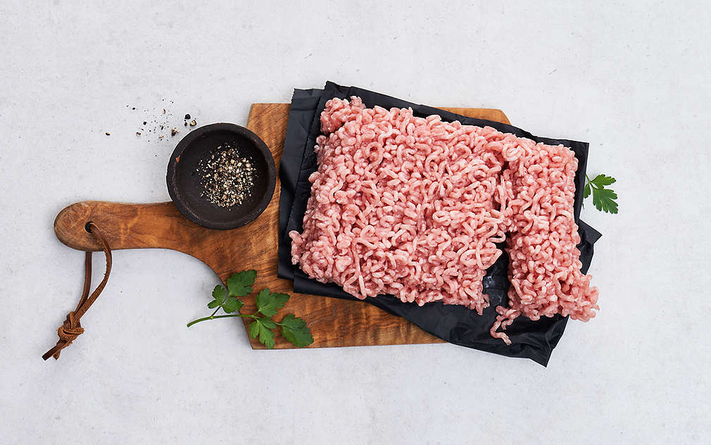 Mljeveno meso na komadu crnog papira na drvenoj dasci. Na drvenoj dasci nalazi se i nekoliko listova peršina i mala zdjelica crnog papra.