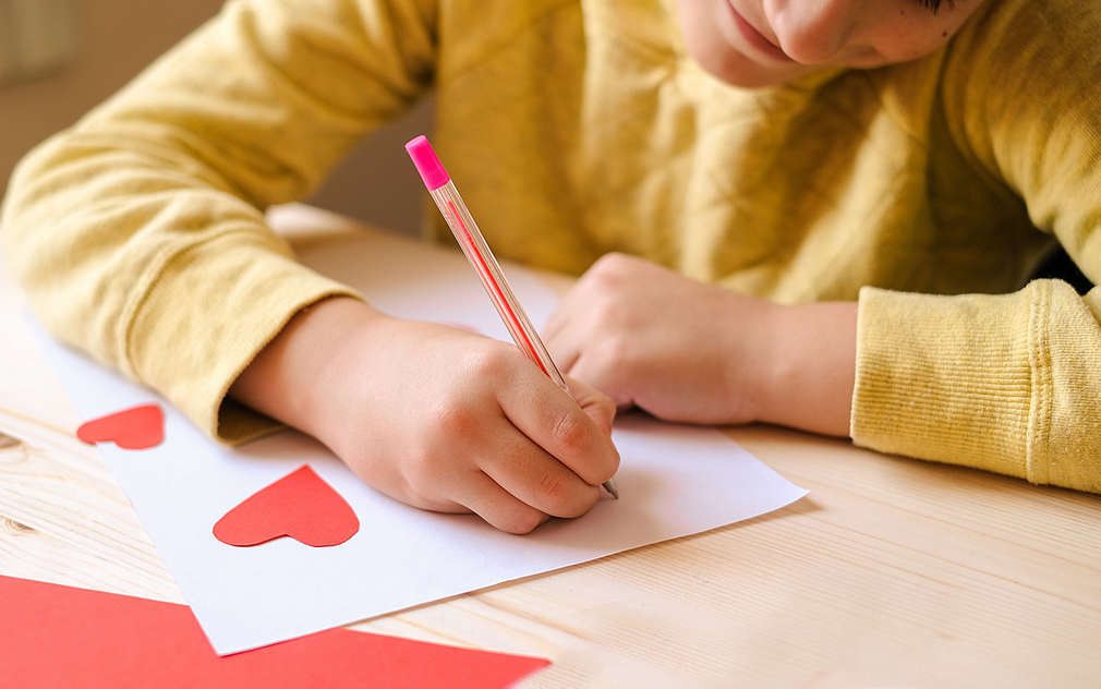 Se poate vedea un băiețel care decorează o felicitare albă cu inimi roșii.