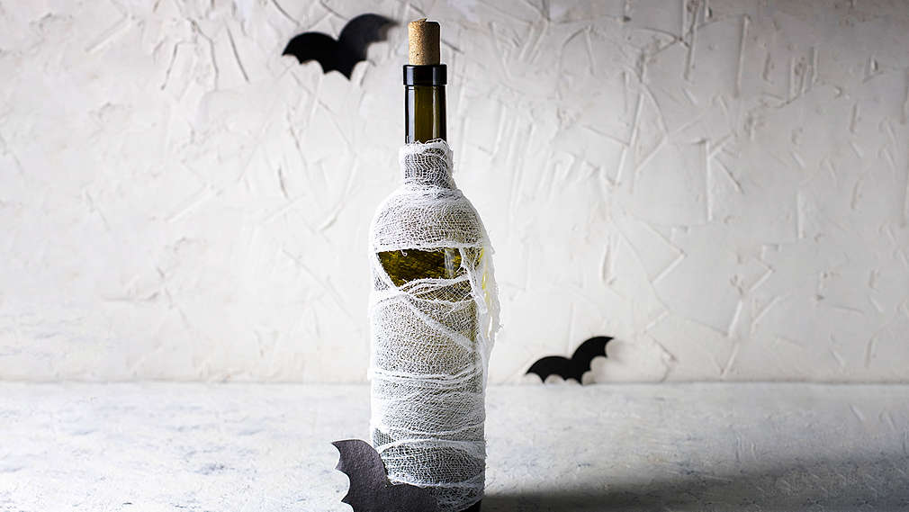 Perete alb structurat în fundal, suprafață gri deschis pe care sunt așezate o sticlă de vin cu dop, înfășurată în tifon. În fotografie mai există trei lilieci mici, decorativi, din hârtie.