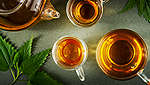 На масата са поставени няколко чаши чай от коприва, а до тях са разположени няколко заснети отгоре листа от коприва.